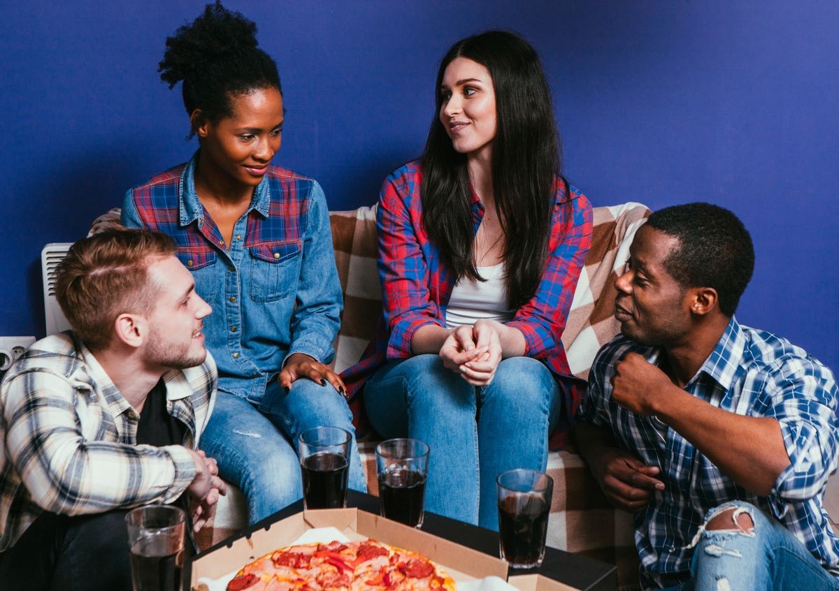 Four young interracial friends sit on couch at home with fresh pizza. Friendship, communication, fun and leisure , relationship, jealousy, gossip concept.